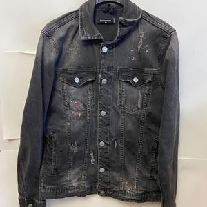 May include: A black denim jacket with a distressed finish, featuring button closures, chest pockets, and a classic collar. The jacket has paint splatter details in red and white. The brand name is visible on the collar.