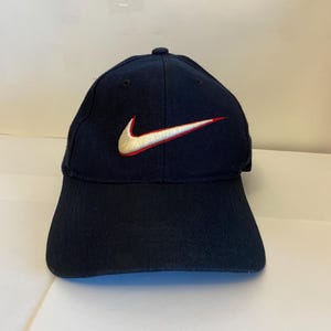 Puede incluir: Una gorra de béisbol azul marino con un logotipo Nike swoosh bordado en el centro. El logotipo es blanco con un fino contorno rojo. La gorra tiene una corona estructurada, una visera curva y ojales cosidos en los paneles superiores.