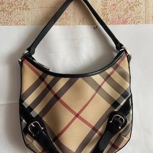 May include: A beige shoulder bag with a classic plaid pattern in red, black, and tan. The bag features black leather trim, a black leather strap, and silver-tone buckles. The bag is a crescent shape.