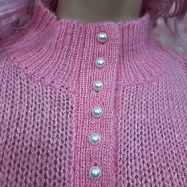 May include: A pink knitted sweater with a row of pearl buttons down the front.
