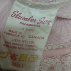 Pode incluir: Um close-up de uma etiqueta de tecido rosa com o texto "Slumber Suzy DREAMWEAR" em script rosa. A etiqueta tamb&eacute;m inclui "MADE IN FABRIQUE AU CANADA" e "100% NYLON". O tamanho "SMALL" &eacute; impresso na parte inferior.