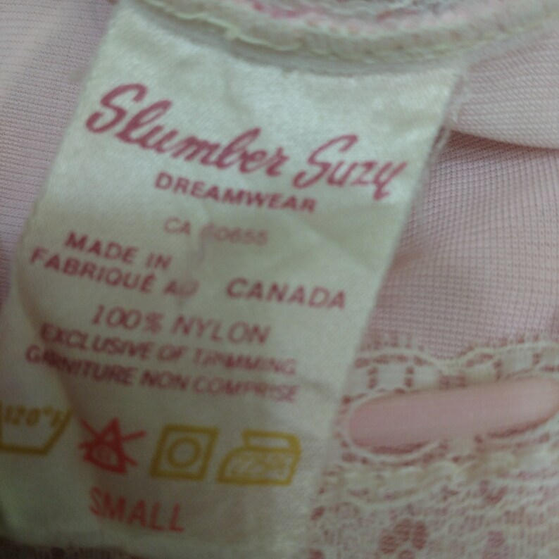 Pode incluir: Um close-up de uma etiqueta de tecido rosa com o texto "Slumber Suzy DREAMWEAR" em script rosa. A etiqueta tamb&eacute;m inclui "MADE IN FABRIQUE AU CANADA" e "100% NYLON". O tamanho "SMALL" &eacute; impresso na parte inferior.