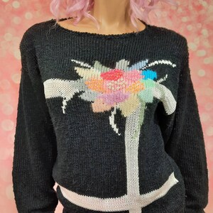 May include: Black knitted sweater with a white and colorful floral design. The sweater has a crew neck and long sleeves.