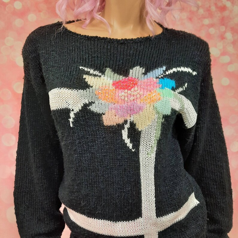 May include: Black knitted sweater with a white and colorful floral design. The sweater has a crew neck and long sleeves.