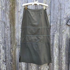 May include: A long, dark green skirt with a straight silhouette and multiple horizontal seams. The skirt is hanging on a wooden hanger against a rustic wooden background.