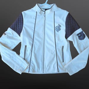 May include: A white jacket with black trim, a stand-up collar, and a full-length zip. The jacket has quilted dark grey sleeves and a logo patch on the upper arm. Two zipped front pockets and a chest logo complete the design.