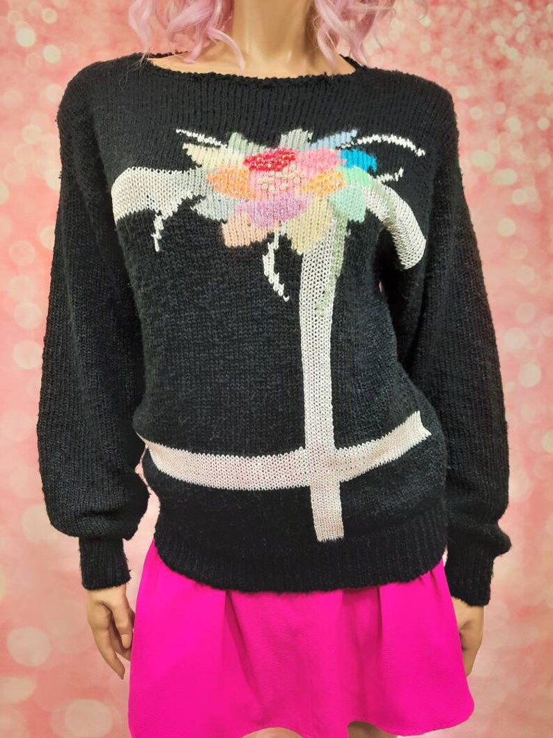 May include: Black sweater with a white and colorful floral design. The sweater has long sleeves and a crew neck.