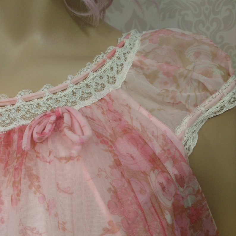 Pode incluir: Camisa de dormir rosa claro com um padr&atilde;o floral. A camisa de dormir apresenta uma delicada renda ao longo do decote e das mangas, com pequenos bot&otilde;es rosa e um la&ccedil;o. As mangas s&atilde;o transparentes e o tecido &eacute; leve.