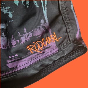 Op de afbeelding: Zwarte boardshorts met een kleurrijke grafische print en de geborduurde oranje tekst "Rip Curl". De shorts hebben een tailleband met trekkoord en zijn gemaakt van lichtgewicht, sneldrogend materiaal.