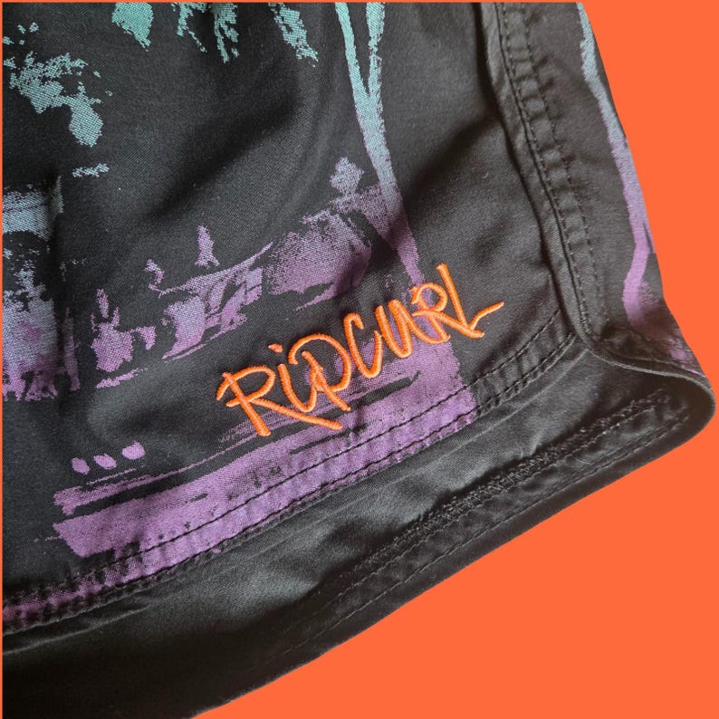 Op de afbeelding: Zwarte boardshorts met een kleurrijke grafische print en de geborduurde oranje tekst "Rip Curl". De shorts hebben een tailleband met trekkoord en zijn gemaakt van lichtgewicht, sneldrogend materiaal.