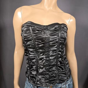 May include: Black strapless corset top with a ruched design. The corset features a row of buttons down the center and vertical ruching details. The top is made of a shiny, satin-like material.