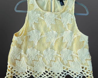May include: A sleeveless, cream-colored top with a floral lace overlay. The top features a round neckline, a zipper closure, and a scalloped hemline. The garment is hanging on a gray hanger.