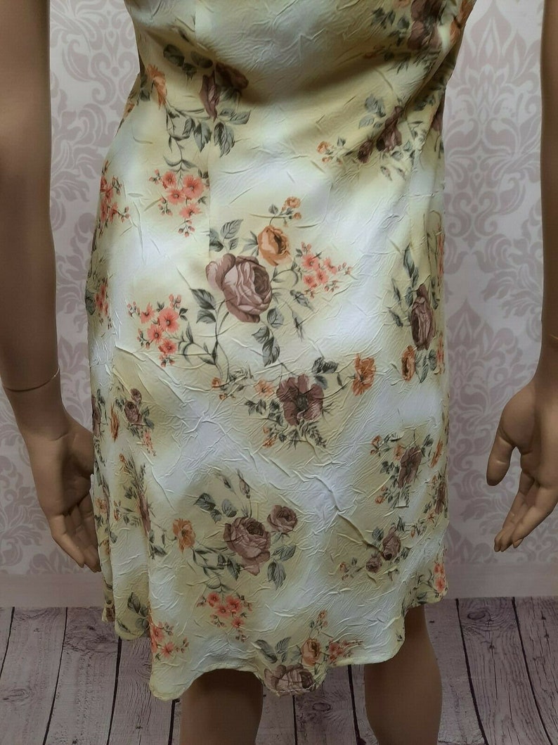 May include: A yellow floral dress with a white background. The dress has a fitted bodice and a flowing skirt. The floral pattern is made up of roses, leaves, and other small flowers.