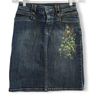 May include: A dark wash denim skirt with a floral sequin design on the right side. The skirt has a classic five-pocket design, a button and zipper closure, and belt loops. The skirt is hanging on a white hanger.
