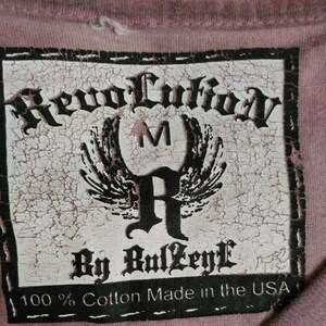 May include: A white tag with black text that reads "Revolution By RulZeyl 100% Cotton Made in the USA". The tag has a distressed look and features a stylized "R" with wings and a "M" above it.