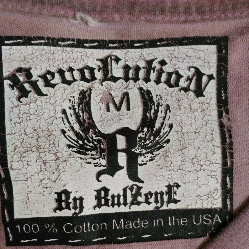 May include: A white tag with black text that reads "Revolution By RulZeyl 100% Cotton Made in the USA". The tag has a distressed look and features a stylized "R" with wings and a "M" above it.