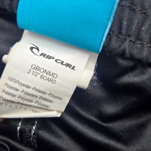 Op de afbeelding: Close-up van een label van Rip Curl boardshorts. Het label is wit met zwarte tekst, inclusief het merklogo en de tekst "GBONMD 2 1/2" BOARD". De shorts zijn zwart met een glanzende textuur en een blauw label is bevestigd.