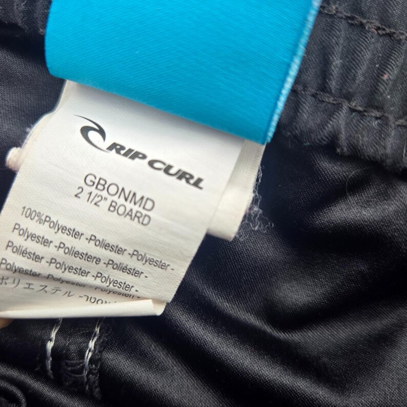 Op de afbeelding: Close-up van een label van Rip Curl boardshorts. Het label is wit met zwarte tekst, inclusief het merklogo en de tekst "GBONMD 2 1/2" BOARD". De shorts zijn zwart met een glanzende textuur en een blauw label is bevestigd.
