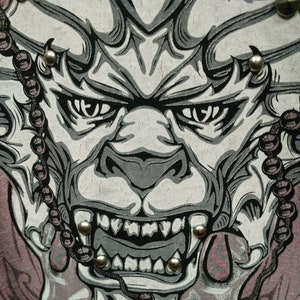 May include: A close-up of a gray and black graphic print of a demon face with sharp teeth and a beaded chain on either side. The demon's eyes are wide open and staring.
