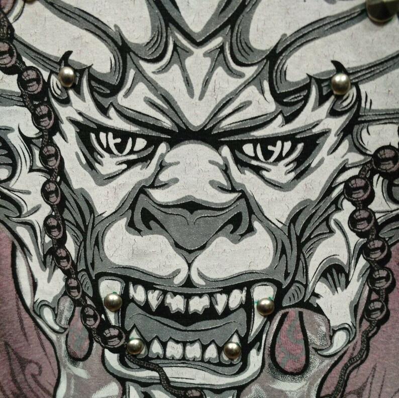 May include: A close-up of a gray and black graphic print of a demon face with sharp teeth and a beaded chain on either side. The demon's eyes are wide open and staring.
