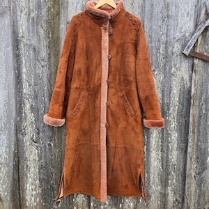 May include: A long brown suede coat with a shearling lining. The coat has a stand-up collar, a button closure, and two pockets. The sleeves have a shearling cuff.
