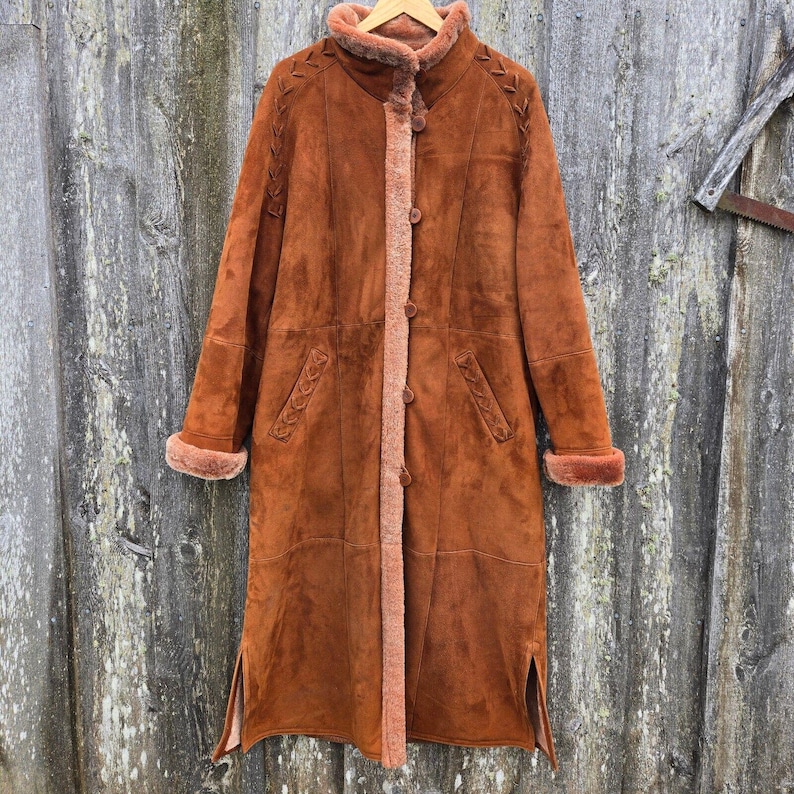 May include: A long brown suede coat with a shearling lining. The coat has a stand-up collar, a button closure, and two pockets. The sleeves have a shearling cuff.