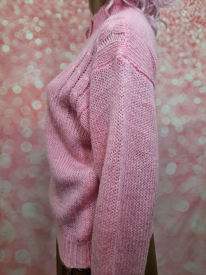 May include: A pink knitted sweater with a cable knit pattern. The sweater has a high neck and long sleeves.