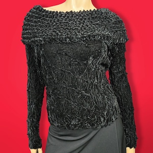 May include: Black long-sleeved shirt with a textured, crinkled fabric. The shirt has a wide, ruffled neckline.