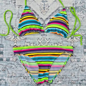 May include: A two-piece bikini set featuring a vibrant horizontal striped pattern in shades of neon green, bright yellow, orange, purple, black, and white. The top consists of two triangular cups with neon green ties, and the bottom is a classic brief.