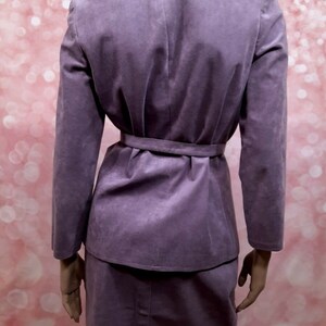 May include: A purple suede skirt suit with a matching jacket and skirt. The jacket has a belt and a stand-up collar.