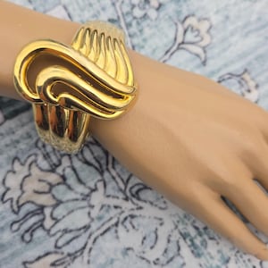 May include: A wide gold-tone cuff bracelet featuring a polished finish and a prominent sculptural design with layered, curved, and ribbed metal elements that create a continuous, flowing pattern across the surface of the wrist accessory.