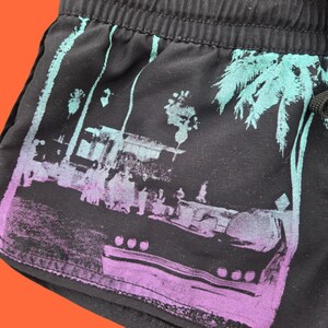 Op de afbeelding: Zwarte short met een tropische sc&egrave;ne print. De print toont palmbomen en een boot in tinten teal en paars. De short heeft een tailleband met trekkoord.