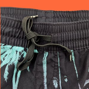 Op de afbeelding: Close-up van een zwarte short met trekkoord en een turquoise palmboomontwerp. De short heeft een zwart trekkoord en een elastische tailleband. De stof heeft een verticaal streepjespatroon.