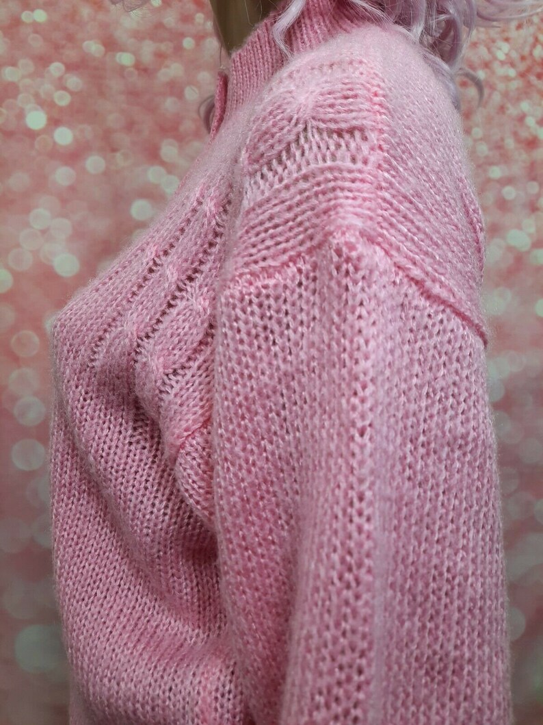 May include: A pink knitted sweater with a cable knit pattern. The sweater has a high neck and long sleeves.