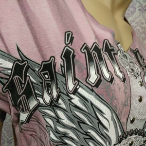 May include: Pink t-shirt with a black and white graphic of an angel wing and the word "Saint" in a bold, gothic font.