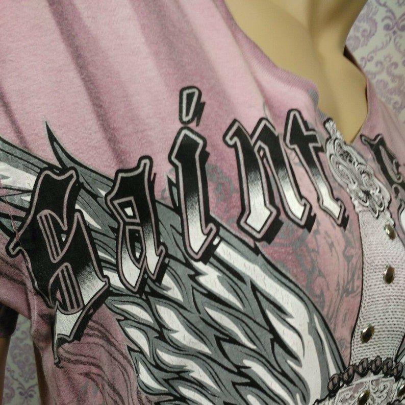 May include: Pink t-shirt with a black and white graphic of an angel wing and the word "Saint" in a bold, gothic font.