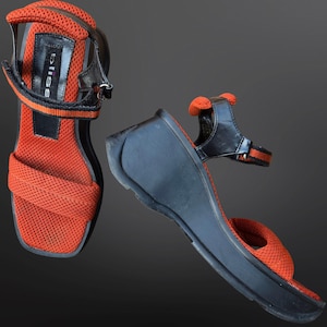 May include: Pair of orange mesh sandals with black wedge heels and straps. The sandals have a square toe design and a strap that wraps around the ankle. The brand name "bliss" is visible on the strap.