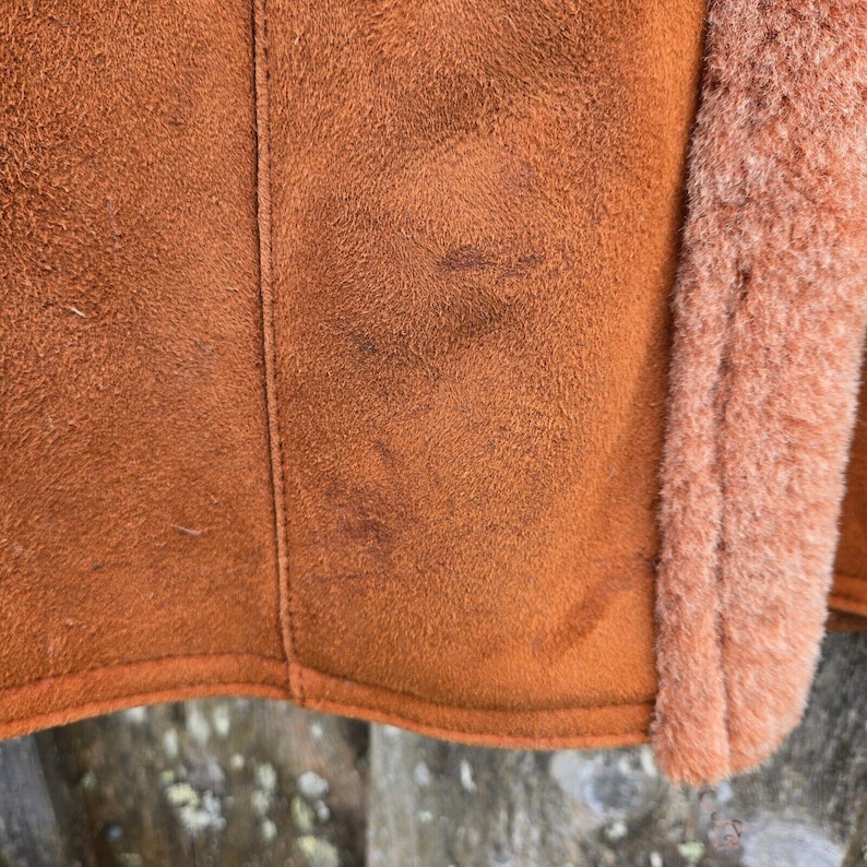 May include: Close-up of a brown suede jacket with a fuzzy orange lining. The suede has a worn look with some light brown marks.