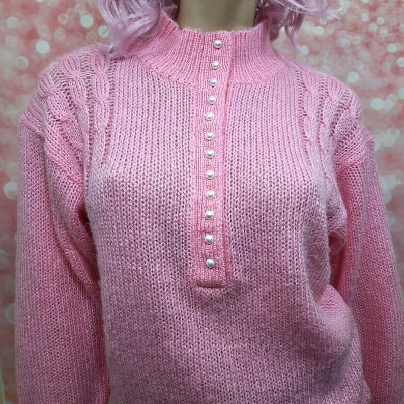 May include: A pink knitted sweater with a mock neck and a row of pearl buttons down the front.