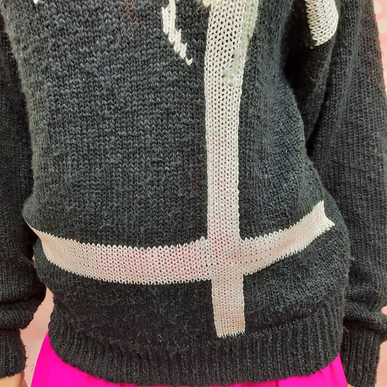 May include: Black knitted sweater with a white knit design of a cross. The sweater is made of a soft, fuzzy material.