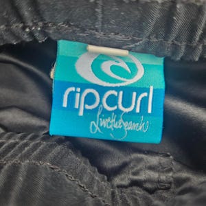 Op de afbeelding: Close-up van een zwart kledingstuk met een Rip Curl-label. Het label is turquoise met de merknaam in het wit en de woorden "Live the Search" in cursief. Het kledingstuk lijkt gemaakt van een glad, mogelijk synthetisch materiaal.