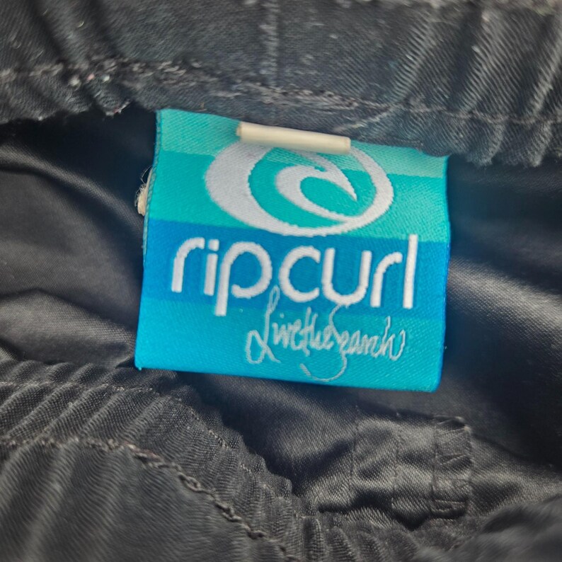 Op de afbeelding: Close-up van een zwart kledingstuk met een Rip Curl-label. Het label is turquoise met de merknaam in het wit en de woorden "Live the Search" in cursief. Het kledingstuk lijkt gemaakt van een glad, mogelijk synthetisch materiaal.