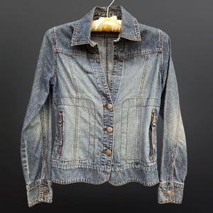 May include: A light blue denim jacket with a button front closure and zippered pockets. The jacket has a classic style with a pointed collar and long sleeves.