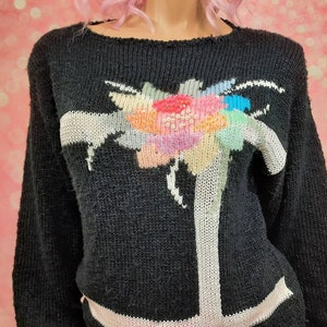 May include: Black knitted sweater with a colorful floral design on the front. The sweater has long sleeves and a crew neck.