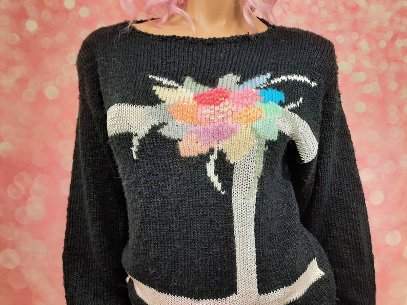May include: Black knitted sweater with a colorful floral design on the front. The sweater has long sleeves and a crew neck.