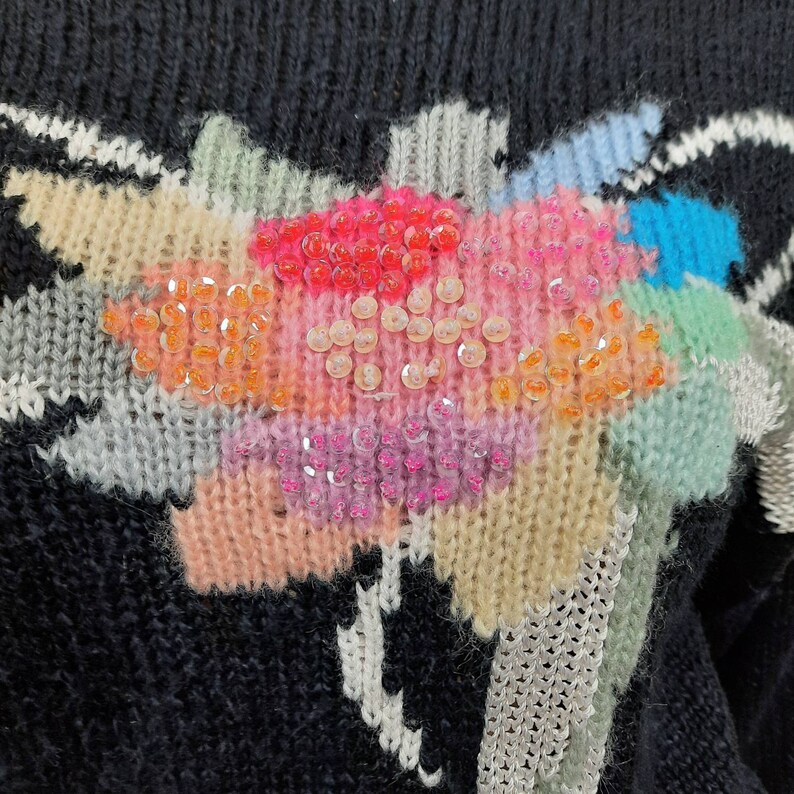 May include: A black knitted sweater with a colorful abstract design featuring sequins. The design includes shades of pink, blue, green, and orange.