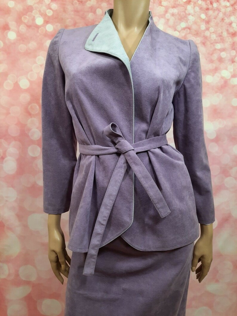 May include: A lavender-colored suede suit with a matching skirt and jacket. The jacket has a contrasting light blue lining and a tie belt.