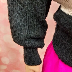 May include: Black knitted sweater with a ribbed cuff. The sweater is partially tucked into a bright pink skirt.