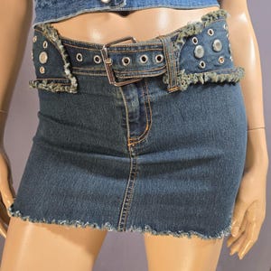 May include: A denim mini skirt with a matching belt. The skirt is a dark wash blue with a frayed hem. The belt has silver grommets and studs, and a silver buckle. The belt is attached to the skirt.