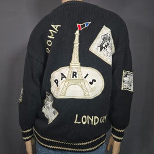 May include: Black knit sweater with embroidered travel-themed patches. Features an Eiffel Tower design with "PARIS" text, and the words "ROMA" and "LONDON". The sweater has a cream-colored trim and a ribbed hem and cuffs.